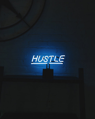Hustle Neon Sign Photowall, tapet cu un semn neon luminos pe un fundal întunecat, ideal pentru a crea o atmosferă modernă și vibrantă.