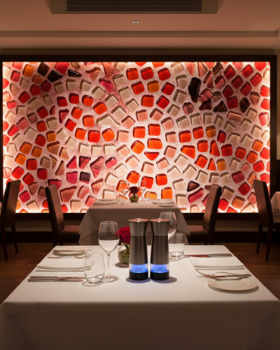 Fototapet personalizat Stained Glass Shards, cu model mozaic de sticlă colorată în nuanțe de roșu, portocaliu și alb, iluminat din spate, decorând peretele unui restaurant elegant cu mese aranjate.