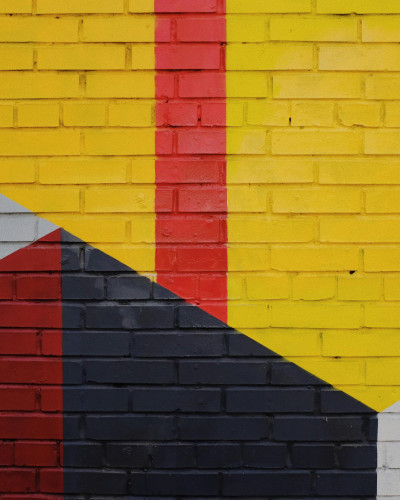 Cameră de tineret cu fototapet modern, design geometric vibrant în culori galben, roșu și gri pe textură de cărămidă. Spațiul include un pat cu lenjerie gri, un puf portocaliu și căști albastre pe o noptieră.
