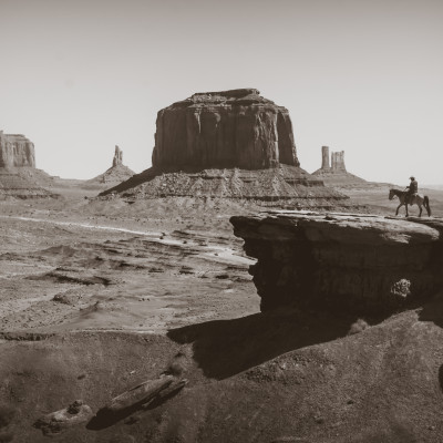 Fototapet personalizat Red Rock Vista, cu un peisaj deșertic vast, formațiuni stâncoase iconice și un cowboy călare, în tonuri sepia. Instalat într-un restaurant modern.